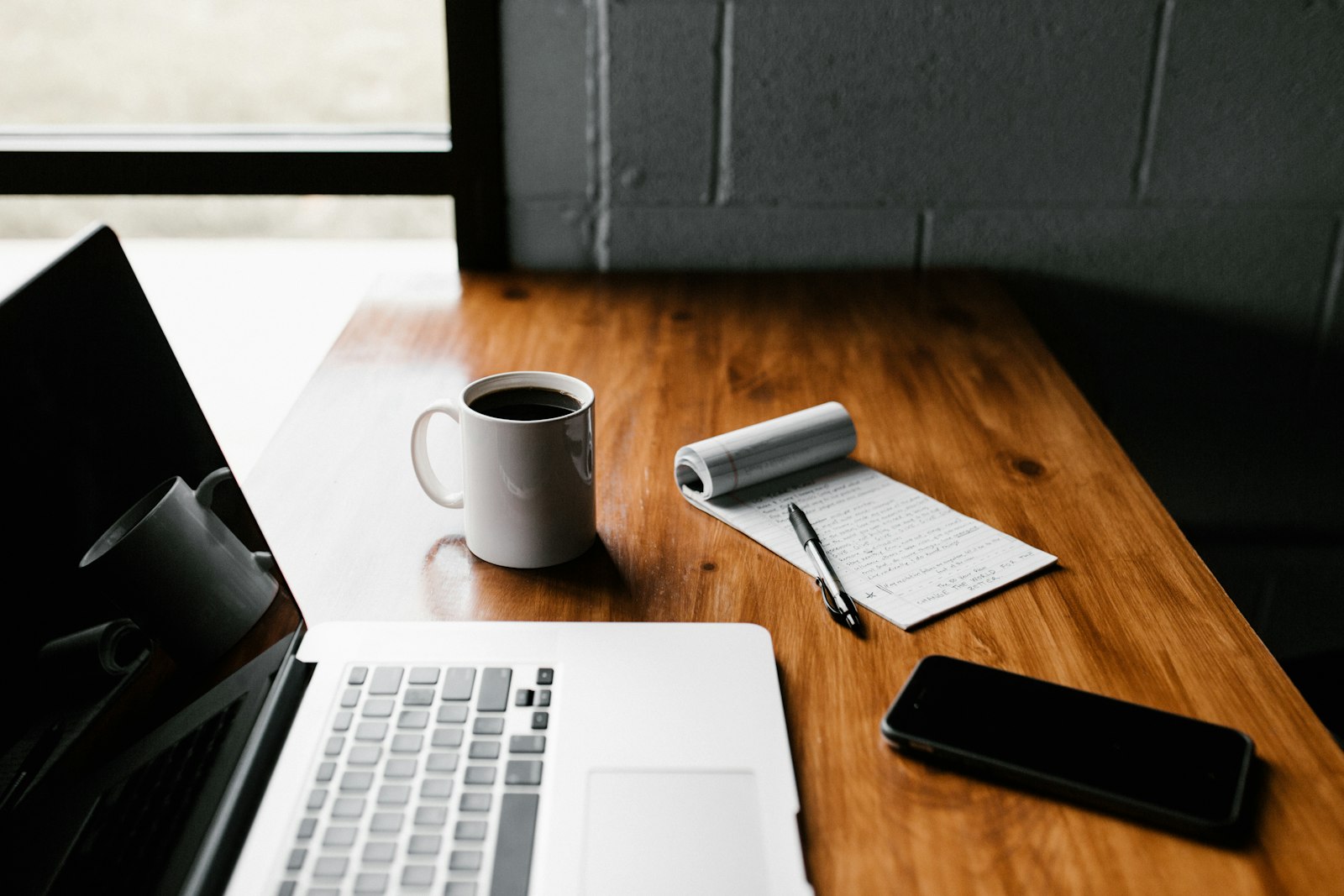Escritorio junto a la ventana con laptop, taza de café y una libreta con notas