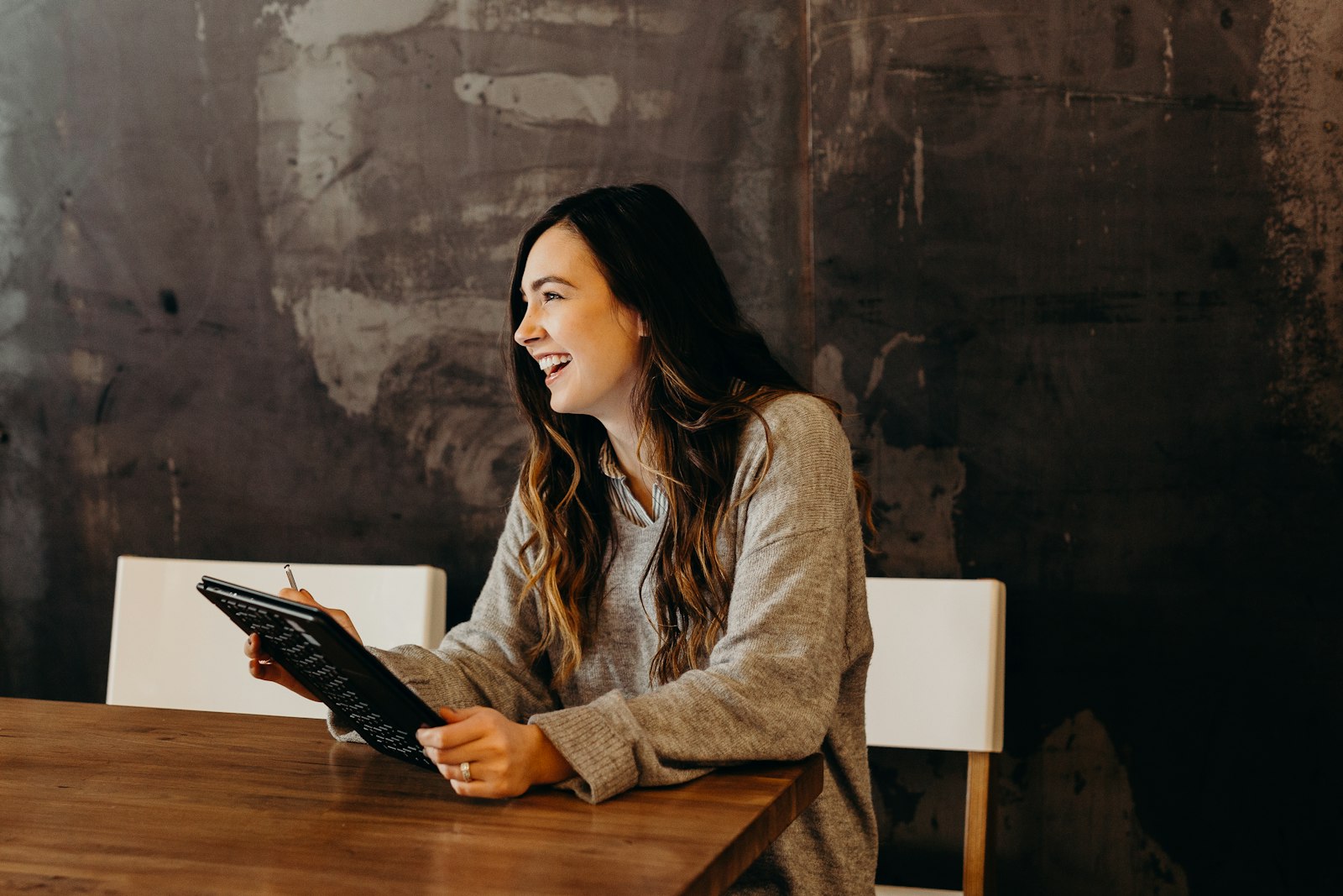 Mujer sonriendo mientras escribe en una tableta sentada a una mesa de madera
